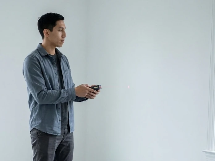 A worker checks an empty white room with a laser measuring tool.